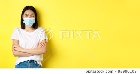 Covid, health care and pandemic concept. Asian woman in white t-shirt and medical mask cross arms on chest and looking sad and disappointed at camera 98996102