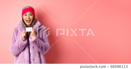 Shopping and fashion concept. Beautiful asian middle-aged woman showing plastic credit card and smiling amazed in stylish clothes, pink background 98996113