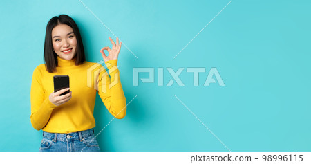 E-commerce and online shopping concept. Portrait of asian woman showing OK sign and using mobile phone, praise app, standing over blue background E-commerce and online shopping concept. Portrait of asian woman showing OK sign and using mobile phone, praise app, standing over blue background 98996115