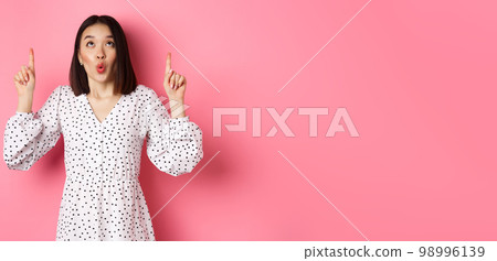 Cute korean girl in beautiful dress saying wow, looking and pointing fingers up, intrigued in promo offer, standing over pink background Cute korean girl in beautiful dress saying wow, looking and pointing fingers up, intrigued in promo offer, standing over pink background 98996139