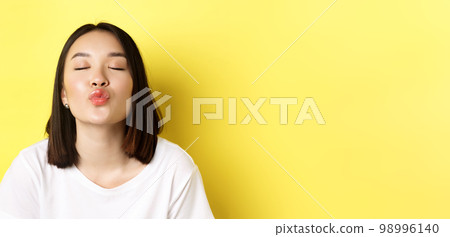 Valentines day concept. Close up of cute asian girl pucker lips and close eyes for kiss, standing romantic against yellow background Valentines day concept. Close up of cute asian girl pucker lips and close eyes for kiss, standing romantic against yellow background 98996140