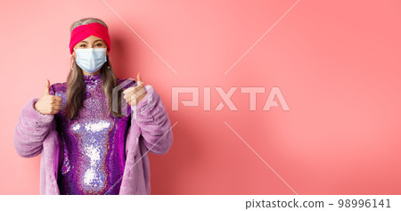 Coronavirus and shopping concept. Stylish asian grandmother in respirator and glittering purple dress and faux fur coat, showing thumbs-up, recommend shop in face masks Coronavirus and shopping concept. Stylish asian grandmother in respirator and glittering purple dress and faux fur coat, showing thumbs-up, recommend shop in face masks 98996141