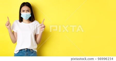 Covid-19, pandemic and social distancing concept. Disappointed asian girl in medical mask, frowning upset and pointing fingers up at logo, standing over yellow background 98996154