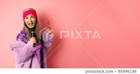 Gorgeous asian senior woman singing karaoke in mic, perform song and looking happy, standing over pink background 98996186