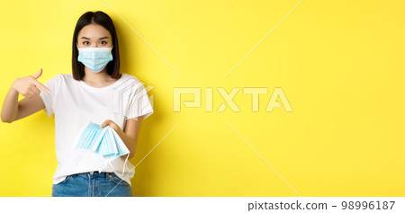 Coronavirus, quarantine and medicine concept. Young asian woman pointing finger at medical masks for going outdoors, standing over yellow background 98996187