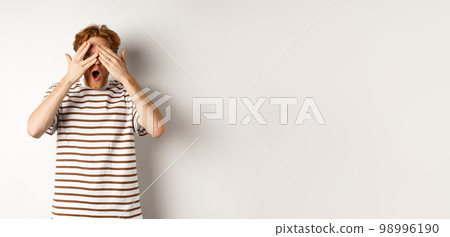 Shocked and embarrassed redhead guy covering eyes, peeking through fingers and gasping, standing over white background 98996190