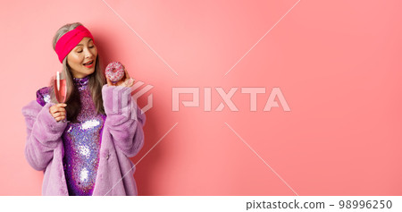 Fancy senior woman having fun, eating donut and drinking pink champagne, standing in purple faux fur coat and glittering dress, studio background 98996250