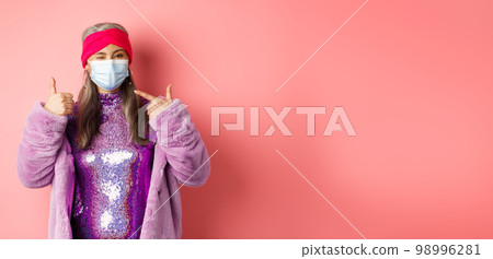Covid-19, social distancing and fashion concept. Smiling asian senior woman in fashionable clothes and respirator, pointing at face mask and show thumb-up, pink background 98996281