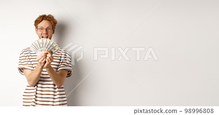Shopping and finance concept. Lucky redhead guy winning, showing prize money and smiling happy, standing in glasses and t-shirt over white background 98996808