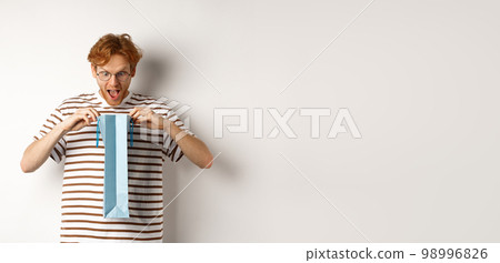 Surprised boyfriend open shopping bag with valentines day gift, staring inside with amazed face, standing over white background Surprised boyfriend open shopping bag with valentines day gift, staring inside with amazed face, standing over white background 98996826