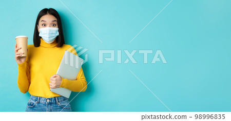 covid-19, health care and quarantine concept. Asian girl student in medical mask standing with laptop and coffee from cafe, standing over blue background covid-19, health care and quarantine concept. Asian girl student in medical mask standing with laptop and coffee from cafe, standing over blue background 98996835