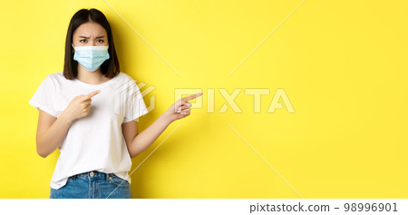 Covid-19, pandemic and social distancing concept. Disappointed asian girl in medical mask, frowning upset and pointing fingers right at logo, standing over yellow background Covid-19, pandemic and social distancing concept. Disappointed asian girl in medical mask, frowning upset and pointing fingers right at logo, standing over yellow background 98996901