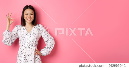Pretty young asian woman in dress showing okay sign, praising and showing approval, looking satisfied, standing against pink background Pretty young asian woman in dress showing okay sign, praising and showing approval, looking satisfied, standing against pink background 98996941