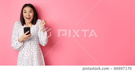 Online shopping and beauty concept. Excited asian woman winning in internet, holding smartphone and rejoicing, smiling happy and celebrating, standing over pink background 98997170