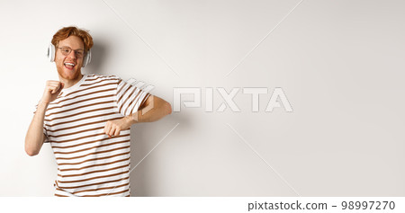 Technology concept. Happy redhead man listening music in headphones and dancing cheerful, standing in t-shirt against white background Technology concept. Happy redhead man listening music in headphones and dancing cheerful, standing in t-shirt against white background 98997270