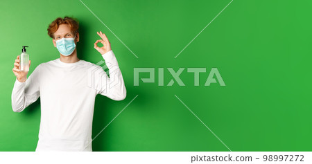 Covid-19, virus and social distancing concept. Smiling young man with red hair, wearing face mask from coronavirus, showing okay sign and hand sanitizer, green background 98997272
