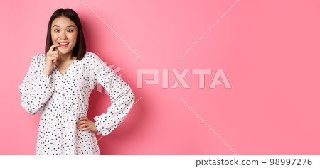Image of beautiful young lady in dress, biting finger and smiling, staring with temptation and amazement, want buy something, standing over pink background 98997276