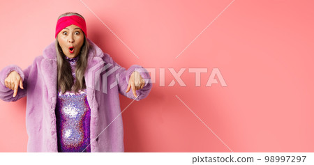Fashion and shopping concept. Impressed asian female pointing fingers down and looking excited at camera, showing awesome promo offer, standing over pink background Fashion and shopping concept. Impressed asian female pointing fingers down and looking excited at camera, showing awesome promo offer, standing over pink background 98997297