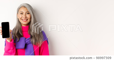 Online shopping. Close-up of smiling asian grandmother showing blank smartphone screen, recommending promotion, standing over white background Online shopping. Close-up of smiling asian grandmother showing blank smartphone screen, recommending promotion, standing over white background 98997309