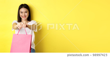 Cute asian girl giving you gift, stretch out hand with pink shopping bag and smiling, congratulating with holiday, standing over yellow background 98997423