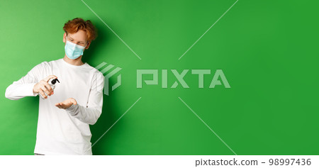 Covid-19, virus and social distancing concept. Redhead guy in medical mask using hand sanitizer and smiling, preventive measures against coronavirus, green background 98997436