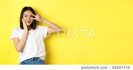 Lovely asian girl in white t-shirt posing with hand on cheek, showing peace sign on eye, standing over yellow background 98997438