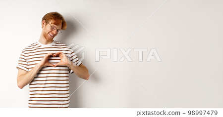 Valentines day. Happy boyfriend with red hair, smiling and showing heart gesture, I love you, standing over white background Valentines day. Happy boyfriend with red hair, smiling and showing heart gesture, I love you, standing over white background 98997479