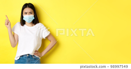 Covid, health care and pandemic concept. Asian female model in medical mask and white t-shirt pointing finger at upper left corner logo, showing promotion, yellow background Covid, health care and pandemic concept. Asian female model in medical mask and white t-shirt pointing finger at upper left corner logo, showing promotion, yellow background 98997549