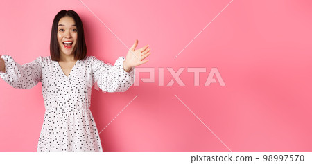 Happy asian girl stretching hands to camera, reaching for hug, smiling and wanting to hold something, standing over pink background in dress Happy asian girl stretching hands to camera, reaching for hug, smiling and wanting to hold something, standing over pink background in dress 98997570