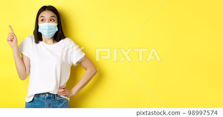 Covid, health care and pandemic concept. Asian woman in medical mask and white t-shirt pointing finger at upper left corner logo, showing promotion, yellow background 98997574