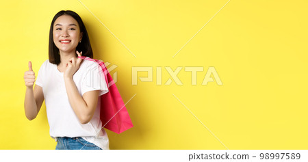Cheerful asian female shopper looking amused, holding shopping bag and showing thumbs up, recommend store with discounts, standing over yellow background 98997589