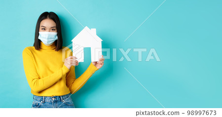 Covid-19, pandemic and real estate concept. Young asian woman in yellow clothes showing paper house cutout, wearing medical mask during coronavirus, standing over blue background Covid-19, pandemic and real estate concept. Young asian woman in yellow clothes showing paper house cutout, wearing medical mask during coronavirus, standing over blue background 98997673