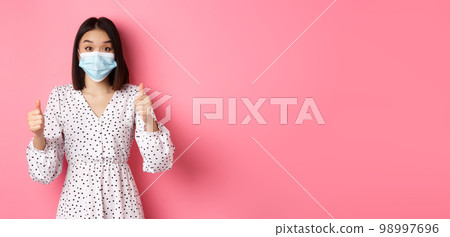 Covid-19, quarantine and lifestyle concept. Excited asian woman in face mask showing thumbs-up, praising good job, looking amazed at camera, standing over pink background Covid-19, quarantine and lifestyle concept. Excited asian woman in face mask showing thumbs-up, praising good job, looking amazed at camera, standing over pink background 98997696