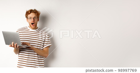 Impressed young man holding laptop in hands, staring at camera with excited face, reading online promotion on website, standing against white backround Impressed young man holding laptop in hands, staring at camera with excited face, reading online promotion on website, standing against white backround 98997769