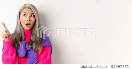 Curious asian senior woman pointing at something interesting, looking at promo wondered, standing in trendy outfit over white background Curious asian senior woman pointing at something interesting, looking at promo wondered, standing in trendy outfit over white background 98997775