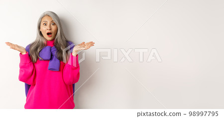 Surprised and amazed asian elderly woman spread hands sideways in complete disbelief, gasping and staring impressed at camera, standing over white background Surprised and amazed asian elderly woman spread hands sideways in complete disbelief, gasping and staring impressed at camera, standing over white background 98997795