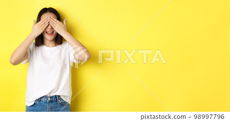 Happy asian woman waiting for surprise gift, close eyes and smiling, expecting something, standing over yellow background Happy asian woman waiting for surprise gift, close eyes and smiling, expecting something, standing over yellow background 98997796
