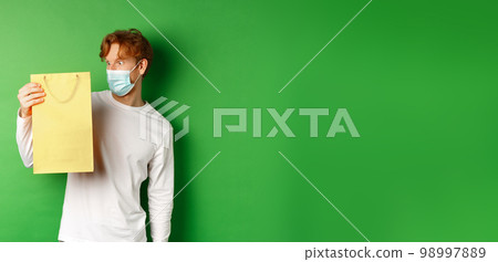 Surprised redhead guy receiving a present, looking at shopping bag with curious face, standing over green background in face mask from covid-19 Surprised redhead guy receiving a present, looking at shopping bag with curious face, standing over green background in face mask from covid-19 98997889