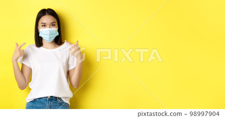 Covid-19, quarantine and social distancing concept. Young asian woman in white t-shirt, pointing at her medical mask from coronavirus, yellow background 98997904
