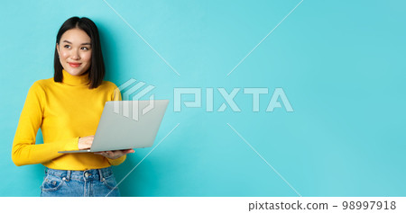 Image of stylish korean woman working on laptop and looking away with dreamy smile, standing with computer against blue background Image of stylish korean woman working on laptop and looking away with dreamy smile, standing with computer against blue background 98997918