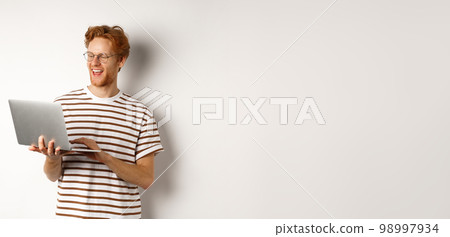 Handsome redhead man working freelance on laptop, smiling and looking pleased at computer screen, white background Handsome redhead man working freelance on laptop, smiling and looking pleased at computer screen, white background 98997934