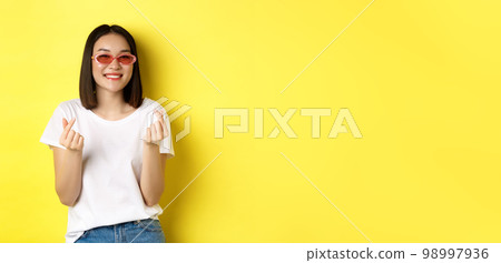 Fashion and lifestyle concept. Attractive asian woman in stylish sunglasses, showing finger hearts and smiling happy at camera, standing over yellow background 98997936