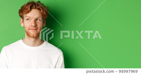 Emotions and fashion concept. Close-up of handsome redhead male model with beard and messy haircut, looking left and smiling, green background 98997969