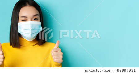 Covid-19, social distancing and pandemic concept. Cute asian girl in medical mask winking at camera, showing thumbs up, good job gesture, praise nice work, blue background 98997981