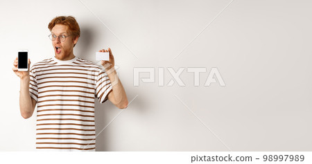 Shopping and finance concept. Amazed young man with red hair showing plastic credit card and smartphone blank screen, staring at display impressed, white background 98997989