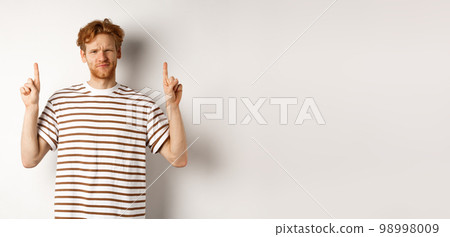 Skeptical handsome redhead man pointing fingers up, grimacing and looking doubtful at camera, standing indecisive over white background Skeptical handsome redhead man pointing fingers up, grimacing and looking doubtful at camera, standing indecisive over white background 98998009