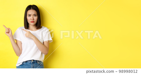 Beauty and fashion concept. Beautiful asian woman in white t-shirt pointing fingers left, standing over yellow background Beauty and fashion concept. Beautiful asian woman in white t-shirt pointing fingers left, standing over yellow background 98998012
