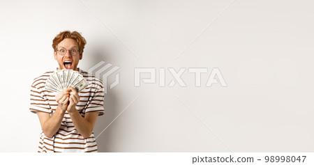 Lucky young man with red hair showing dollars, winning money and screaming of happiness, holding prize cash, standing over white background Lucky young man with red hair showing dollars, winning money and screaming of happiness, holding prize cash, standing over white background 98998047
