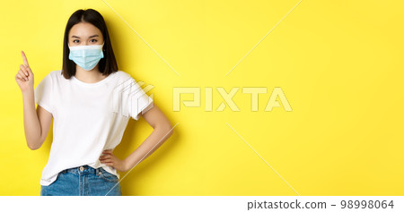 Covid, health care and pandemic concept. Asian female model in medical mask and white t-shirt pointing finger at upper left corner logo, showing promotion, yellow background 98998064