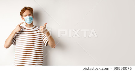 Covid-19, pandemic and social distancing concept. Cheerful young man with red hair pointing at medical mask and showing thumb-up, recommend use preventive measures against virus Covid-19, pandemic and social distancing concept. Cheerful young man with red hair pointing at medical mask and showing thumb-up, recommend use preventive measures against virus 98998076